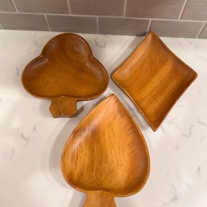 May include: Three wooden serving dishes in the shapes of playing card suits: a club, a diamond, and a spade. The dishes are made of light brown wood and have a smooth, polished finish. The club and spade dishes have stems.