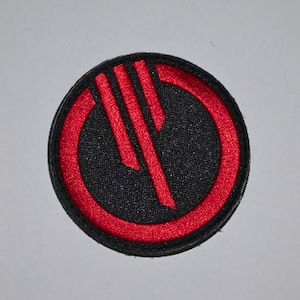 May include: A red and black embroidered patch with a circular design. The circle has a red outline and a black interior with a red stylised symbol in the centre.