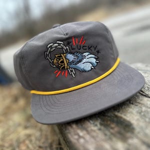 May include: Gray baseball cap with a yellow rope trim and an embroidered paw print with a crown and chain. The paw print is blue and the crown is gold. The text "LUCKY" is embroidered in black above the paw print.