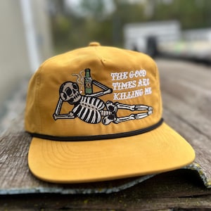 May include: Mustard yellow cap with a black rope detail and a flat brim. The cap features an embroidered skeleton graphic holding a beer bottle, with the text "THE GOOD TIMES ARE KILLING ME".
