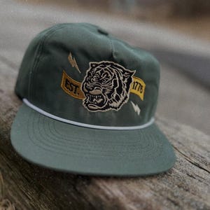 Op de afbeelding: Een groene baseballcap met een witte rand en een geborduurde tijgerkop aan de voorkant. De tijger heeft een geel en zwart streepdessin en de tekst "EST. 1776" is geborduurd op de cap.