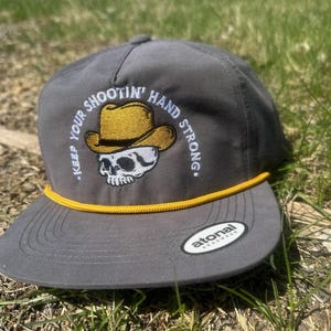 Könnte beinhalten: Graue Baseballkappe mit gelber Kordel und einem Totenkopf, der einen gelben Cowboyhut trägt, der auf der Vorderseite gestickt ist. Der Text "Keep Your Shootin' Hand Strong." ist um den Totenkopf herum gestickt. Die Kappe hat ein schwarz-weißes "Atonal"-Etikett an der Seite.