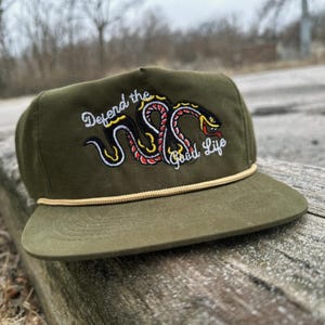Puede incluir: Una gorra de béisbol verde con ribete de cuerda y una serpiente bordada con el texto "Defend the Good Life".