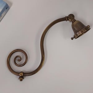 May include: An antique brass-toned wall-mounted reading lamp with a bell-shaped shade. The lamp features a curved arm with a decorative spiral and a detailed floral design on the shade's top. A book is partially visible in the background.
