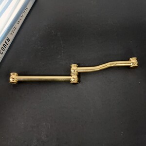 May include: A gold-colored, articulated cabinet pull with a distinctive design. The pull features a central joint and two extending arms, each ending in a decorative knob. The hardware is set against a dark background.