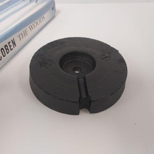 May include: A black, circular weight with a central hole and a slot cut into the side. The weight has a textured surface and appears to be made of cast iron. The weight is next to a book titled "THE WOODS".