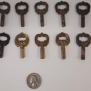 May include: An assortment of antique skeleton keys in various shades of bronze and brass. The keys have ornate, decorative handles and are arranged on a white surface. A quarter is included for size reference.