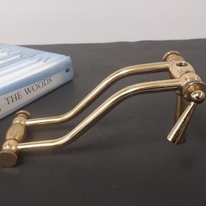 May include: A brass-colored, curved metal object with a handle and adjustable knobs. The object is positioned on a dark surface next to a book titled "The Woods". The metal has a polished finish, reflecting light.