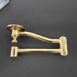 May include: A brass-colored, vintage-style door handle with a curved arm and a circular base. The handle has a rectangular section and a decorative, tiered design. The hardware is likely for a cabinet or door.