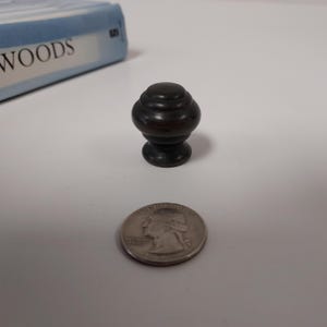 May include: A dark brown, decorative cabinet knob with a layered design. The knob is next to a U.S. quarter for size comparison. The knob is approximately 1 inch in height. A book with the word "WOODS" is in the background.