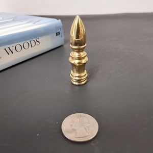 Puede incluir: Un remate de lámpara de latón con una parte superior puntiaguda y detalles decorativos. El remate mide aproximadamente 6,35 cm de alto. Una moneda de veinticinco centavos está al lado del remate para comparar el tamaño. Un libro con el título "WOODS" está en el fondo.
