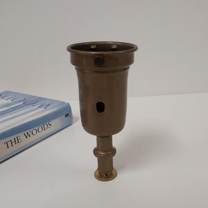 May include: A bronze-colored lamp socket with a cylindrical body and a flared top. It has a small hole and a threaded base. A book titled "THE WOODS" is partially visible in the background.