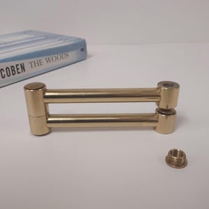 May include: A gold-colored, double-bar metal object with threaded ends and a separate threaded piece. The object is lying on a white surface. A book titled "THE WOODS" is visible in the background.