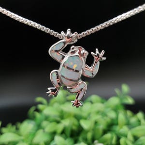 Puede incluir: Un collar de cadena de plata con un colgante de rana de ópalo blanco. La rana está en una pose de escalada con las patas extendidas.