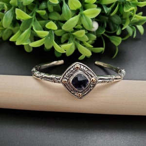 May include: Silver and gold tone bracelet with a square-shaped amethyst gemstone in the center. The bracelet has a woven design and is accented with small gold beads.