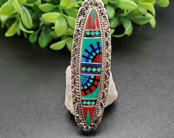 Op de afbeelding: Een zilveren ring met een ovale inleg van turquoise, rode en zwarte stenen. De stenen zijn gerangschikt in een geometrisch patroon.