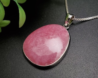 May include: A pink gemstone pendant with a silver-colored bezel and chain. The oval-shaped stone has a textured appearance with a lighter, white area in the center. The necklace is displayed against a dark background.