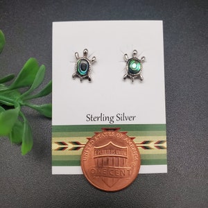 May include: A pair of sterling silver turtle stud earrings with abalone shell inlays. The earrings are displayed on a white card with the text "Sterling Silver". A penny is placed in front of the card.