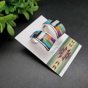 May include: A pair of sterling silver hoop earrings with a geometric inlay of turquoise, orange, purple, and black stones. The earrings are displayed on a white card with the text "Sterling Silver".