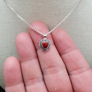 May include: A silver chain necklace with a small red heart pendant. The heart pendant has a silver border with a braided design.