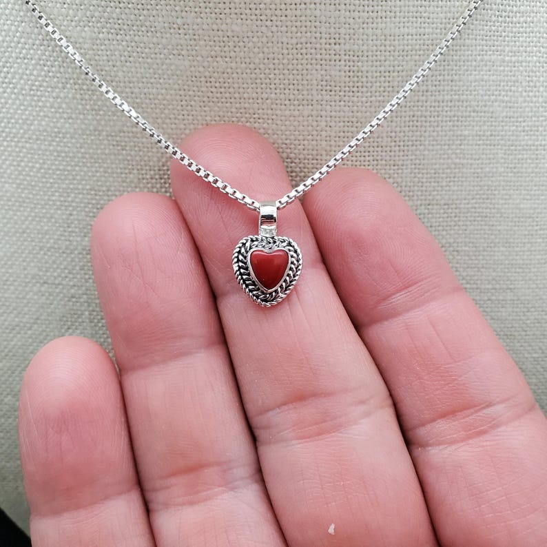 May include: A silver chain necklace with a small red heart pendant. The heart pendant has a silver border with a braided design.