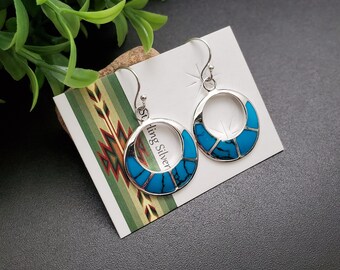 May include: A pair of silver hoop earrings with turquoise inlay. The earrings are circular with a polished silver frame and feature geometric turquoise segments. The earrings are displayed on a white card with the words "Sterling Silver".