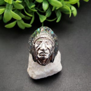 May include: A silver ring with a detailed depiction of a Native American chief wearing a headdress with iridescent turquoise and green stones.