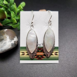 May include: A pair of silver earrings with white mother of pearl teardrop shaped stones. The earrings have a detailed silver design around the stones.