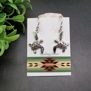 May include: Sterling silver earrings featuring a sea turtle design with abalone shell accents. The earrings are displayed on a card with the text "Sterling Silver".