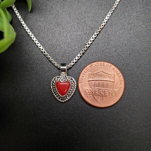 May include: A silver chain necklace with a red heart pendant. The pendant is detailed with silver filigree and a red stone in the center. The necklace is shown next to a US penny for size comparison.