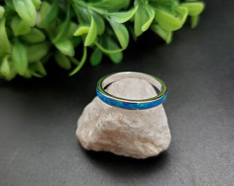 Pode incluir: Um anel de banda de prata com uma fina incrustação de opala azul. O anel está sentado em uma pequena rocha branca.