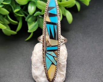 Op de afbeelding: Een zilveren ring met een lange, ovale steen die in het midden is gezet. De steen is gemaakt van turquoise, zwart en wit inlegwerk. De ring heeft een touwachtig ontwerp rond de rand.
