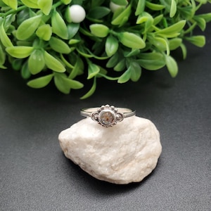 May include: A silver ring with a clear gemstone set in a decorative bezel. The ring is on a white rock.