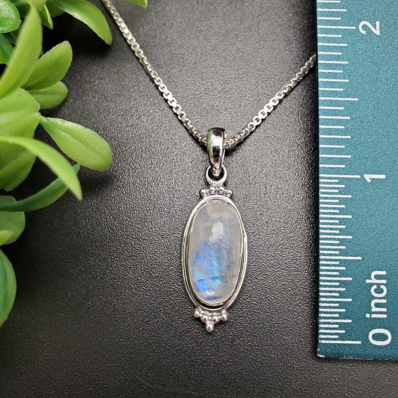 May include: A silver chain necklace with a silver oval pendant featuring a blue and white moonstone. The pendant is approximately 1 inch long.