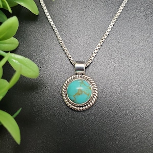 May include: A silver chain necklace with a turquoise stone pendant. The pendant is round and has a silver border.
