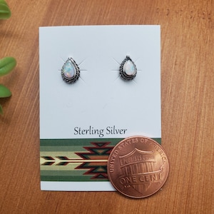 Può includere: Un paio di orecchini in argento sterling con pietre di opale bianco a forma di lacrima. Gli orecchini sono esposti su una carta bianca con le parole "Sterling Silver" stampate su di essa. Un centesimo è incluso come riferimento per le dimensioni.
