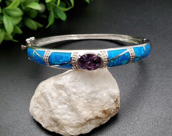 May include: A silver bangle bracelet with a blue opal inlay and a purple amethyst gemstone in the center.
