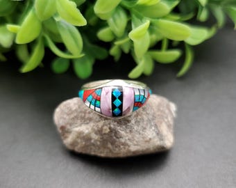 May include: A silver ring with a pink, blue, red, and black inlay design. The ring is set on a gray rock.