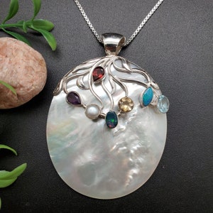May include: A silver pendant necklace with a large white mother-of-pearl oval charm. The charm features a silver tree design with seven gemstones: amethyst, garnet, opal, pearl, citrine, aquamarine, and black onyx.