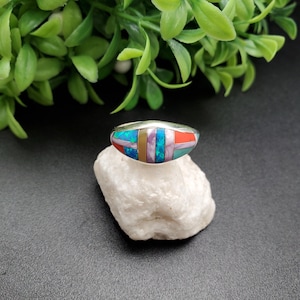 May include: A silver ring with a colorful inlay of turquoise, coral, opal, and purple stones. The ring is set on a white rock.