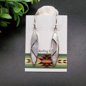 May include: A pair of sterling silver earrings with a white mother-of-pearl inlay. The earrings are shaped like a teardrop and have a geometric design. The earrings are on a white card with the text "Sterling S".