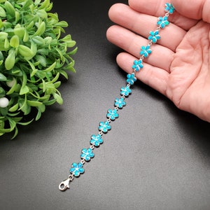 May include: A silver bracelet with a chain of blue flower charms. The bracelet is on a hand, with the hand's fingers spread out.