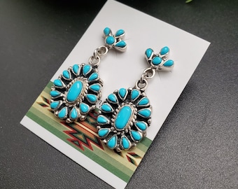 May include: A pair of silver earrings featuring turquoise stones. The earrings are shaped like a flower with a central oval stone surrounded by smaller stones.