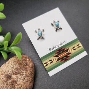 May include: A pair of sterling silver turtle earrings with blue turquoise stones. The earrings are displayed on a white card with the words "Sterling Silver" printed on it.