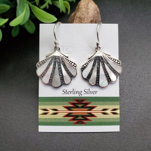 May include: A pair of sterling silver seashell earrings with white mother-of-pearl inlays. The earrings are displayed on a white card with the words "Sterling Silver" printed on it. The card has a green and red geometric design at the bottom.