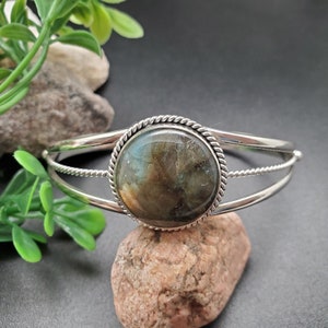 Op de afbeelding: Een zilveren armband met een ronde, iriserende labradorit edelsteen gezet in een zilveren bezel. De armband heeft een delicaat, gedraaid draadontwerp.