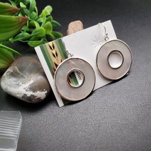 Puede incluir: Un par de pendientes de plata con discos redondos de madreperla blanca. Cada disco tiene un círculo más pequeño cortado en el centro. Los pendientes se muestran en una tarjeta blanca con la palabra "plata" impresa en ella.