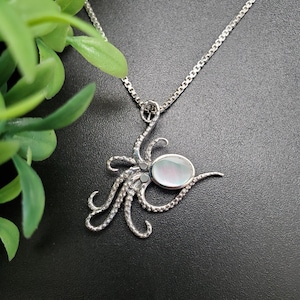 May include: A silver-toned octopus pendant necklace with a pearlescent oval center stone. The octopus design features detailed tentacles and is suspended from a delicate chain. The necklace is set against a dark background with green foliage in the upper left corner.