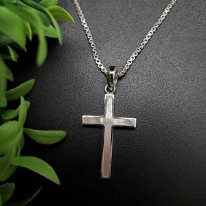 May include: A silver cross pendant necklace. The cross is a simple, polished design with a rectangular shape. The necklace chain is also silver and has a delicate, linked pattern. The background is a dark, solid color, with green leaves in the upper left corner.
