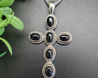 Puede incluir: Un colgante de plata en forma de cruz con cinco piedras ovaladas negras. El colgante está en una cadena de plata.